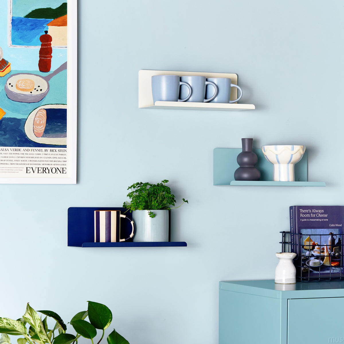 A styled interior scene with light blue walls, floating shelves, a matching blue cabinet, a white table, and a potted green plant. No person or animal is present, but this is not a clean isolated product shot; it shows the product in a real room setting, so subject_type is Lifestyle.